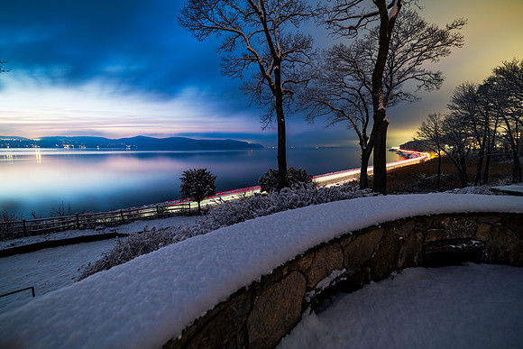 Hudson River after sunset and blue hour following a snow storm with mountains in the background and light trails from the commuter train in the foreground