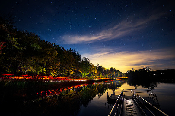 Long Exposure photograph of a road cutting through Fahnestock State Park and pond with the night sky , stars and tail light trails from cars